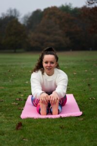 yoga for arthritis teacher, Paulina, smiling directly into the camera while in a park in a forward fold position holding